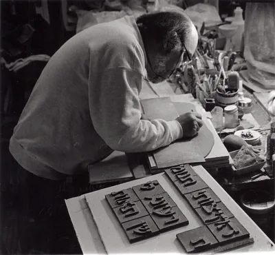 Black and white photograph of Arnold bent over a workbench, cutting large Hebrew letters from slabs of clay