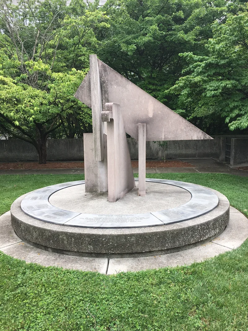 An angular eagle of interlocking pink marble slabs on a round stone base with inscribed panels, in a sunken garden with trees behind