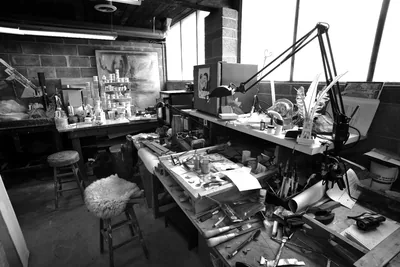Black and white photograph of the enameling bench in the studio, with tools, jars, and a lamp