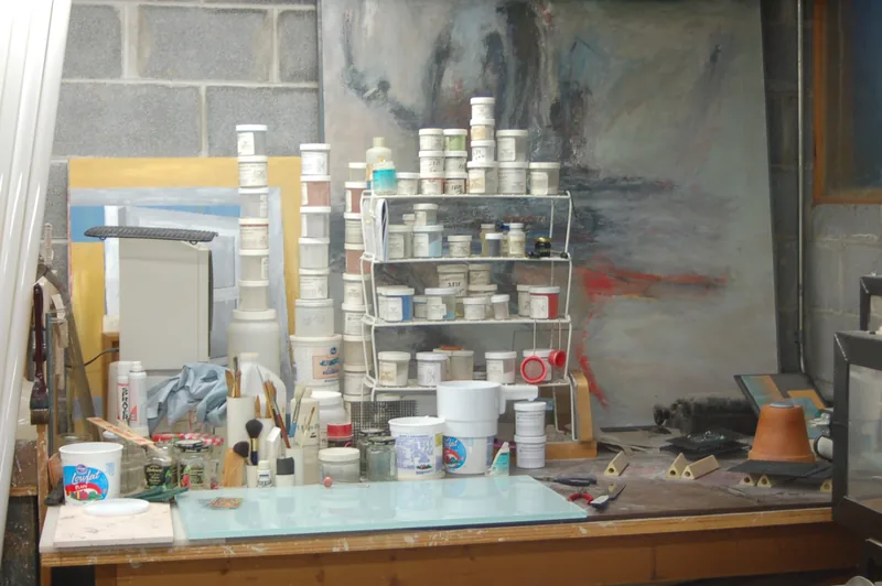 A studio workbench crowded with jars of glaze, brushes, and tools, a large abstract painting behind