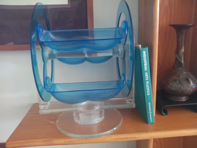 A two-tier lazy susan of blue and clear Plexiglas, two scalloped trays on a clear axle between two blue disks, turning on a round clear base, beside the textbook Industrial Arts Plastics